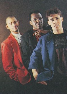 three men standing next to each other in front of a black background with one man wearing a red jacket