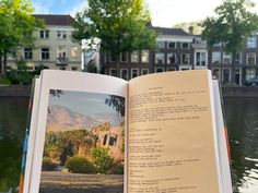 an open book sitting on top of a wooden table next to water and buildings in the background