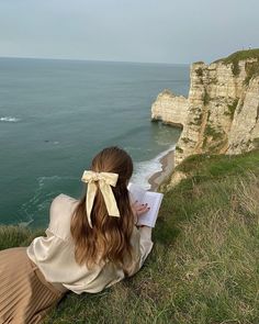 a woman sitting on top of a grass covered hillside next to the ocean reading a book