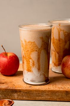 two glasses filled with liquid sitting next to an apple on a wooden tray and one glass has something brown in it