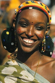 an african woman with painted face and headdress smiles at the camera while standing in front of a crowd