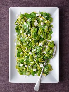 a white plate topped with green vegetables and feta cheese next to a spoon on a purple table cloth