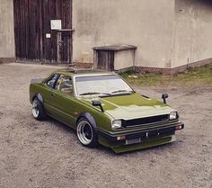 a green car parked on top of a dirt field next to an old building in the background