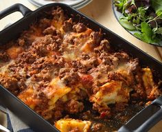 a casserole dish with meat and cheese in it sitting on a table next to a salad