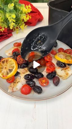 a white plate topped with fish covered in olives, tomatoes and lemon wedges