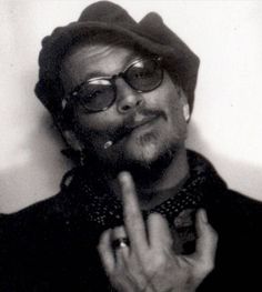 a man wearing glasses and a hat making the peace sign with his hand while standing in front of a white wall