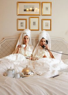 two women in white robes are sitting on a bed with tea cups and telephones