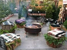 an outdoor fire pit surrounded by potted plants and benches with blankets on them,