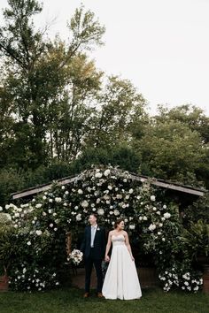 a bride and groom standing in front of an arch covered with white flowers at their wedding