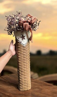 a hand holding a vase with flowers in it on top of a wooden table at sunset