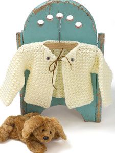 a teddy bear laying next to a crocheted baby sweater on a wooden chair