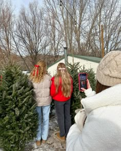 two girls are looking at christmas trees in the snow with their cell phones and one girl is wearing a red jacket