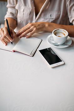 a woman sitting at a table writing in a notebook with a cup of coffee next to her