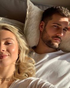 a man and woman laying in bed with pillows on their backs looking at the camera