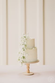 a wedding cake with white flowers on top is sitting on a small wooden stand in front of the wall