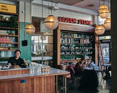 people sitting at tables in a restaurant with lights hanging from the ceiling and shelves full of liquor bottles