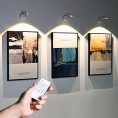 a hand holding a remote control in front of three pictures on the wall with lights