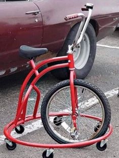 a red bicycle parked next to a maroon car in a parking lot with no wheels