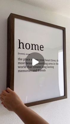 a person holding up a framed poster with the words home on it and a hand pointing at it