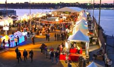 people are walking around an outdoor market by the water at night with lights on them