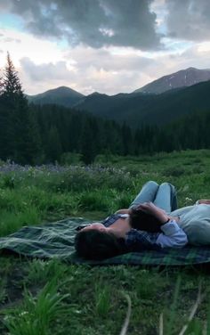 two people laying on a blanket in the middle of a field with mountains in the background