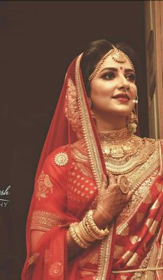 a woman in a red and gold bridal outfit standing next to a window with her hands on her chest