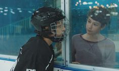 a young boy wearing a hockey helmet looking out the window at another person behind him