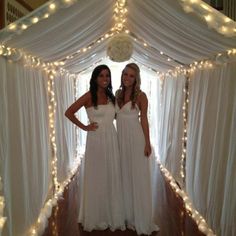 two beautiful women standing in front of a white tent with fairy lights on the ceiling