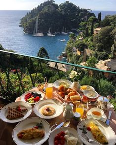 an outdoor table with food and drinks on it overlooking the water's edge in front of some boats