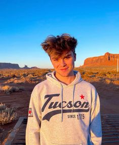 a young man standing in the desert wearing a grey hoodie and smiling at the camera