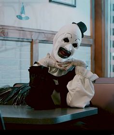 a creepy clown sitting on top of a table next to a black bag and wearing a white mask