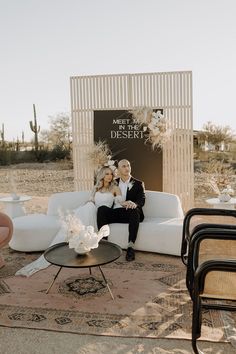 a man and woman sitting on a couch in front of a desert backdrop with chairs