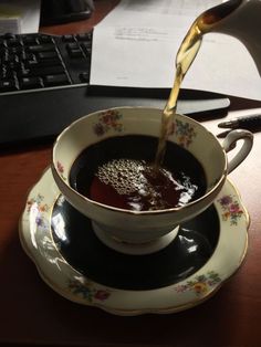 a cup of coffee is being poured into the teacup with saucer and keyboard in the background