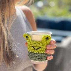 a woman holding a coffee cup with a crocheted frog face on the side