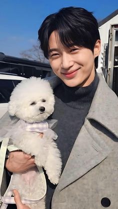 a young man holding a white dog in his hands and smiling at the camera while wearing a gray coat