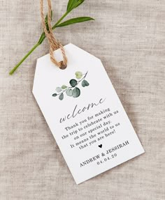 a wedding welcome card hanging from a string on a piece of cloth with a plant in the background