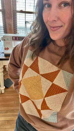 a woman standing in front of a window wearing a brown shirt and jeans with an orange patchwork design on the chest