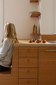 a woman sitting at a kitchen counter with apples on the counter and shelves behind her