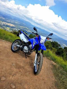 a blue and white motorcycle parked on top of a dirt road next to a lush green hillside