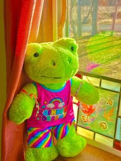 a green teddy bear wearing a pink shirt and rainbow pants leaning against a window sill