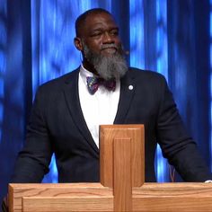 a man with a beard and bow tie standing at a podium in front of a blue curtain