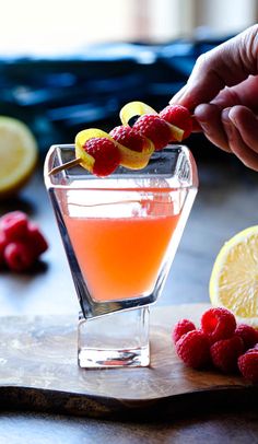 a person holding a piece of fruit over a glass filled with orange juice and raspberries