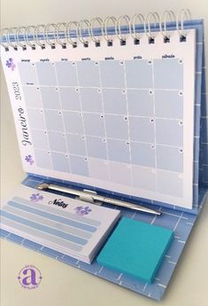 a desk calendar and pen sitting on top of a blue binder next to a notepad