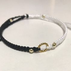 a white and black cord bracelet with two gold beads on it's end, sitting on a table