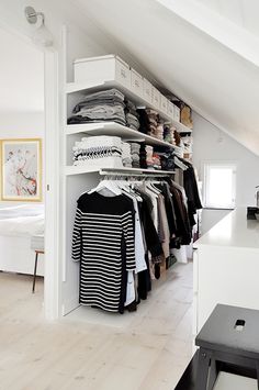 an attic bedroom with white walls and open shelving, clothes on hangers in the closet