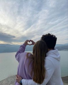 a man and woman sitting on the edge of a cliff with their hands in the shape of a heart