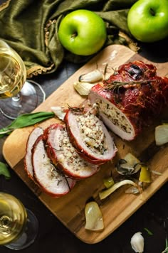 sliced meat and vegetables on a cutting board next to wine glasses, apples and napkins