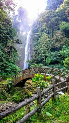 a wooden bridge over a small stream in front of a tall waterfall with a large waterfall coming out of it