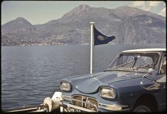 an old car parked on the side of a lake next to a flag pole with mountains in the background