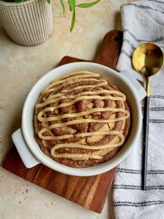 a cinnamon roll in a white bowl on a cutting board with a spoon next to it